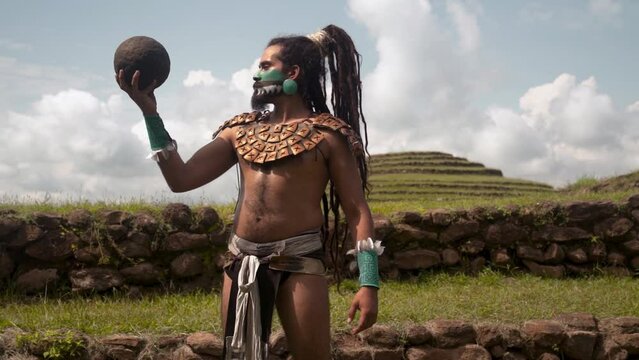 Mayan male warrior in traditional body costumes and face paint engaging in the ancient Mesoamerican ballgame with a dark ball standing in a grass near ancient ruins in Mexico