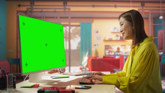 Beautiful Professional Asian Woman Works on Her Personal Computer with Big Green Screen Mock Up Display. Creative Female Works in a Cool Loft Space. Studying and Occupation Concept