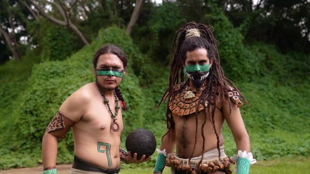 Mayan male warriors in traditional body costumes and face paint engaging in the ancient Mesoamerican ballgame with a dark ball standing looking at camera in a grass near ancient ruins in Mexico