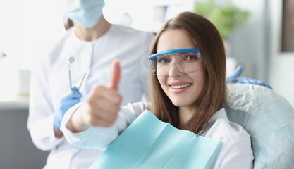 Young woman in safety glasses sitting in dental chair and showing thumbs up. Quality dental care...