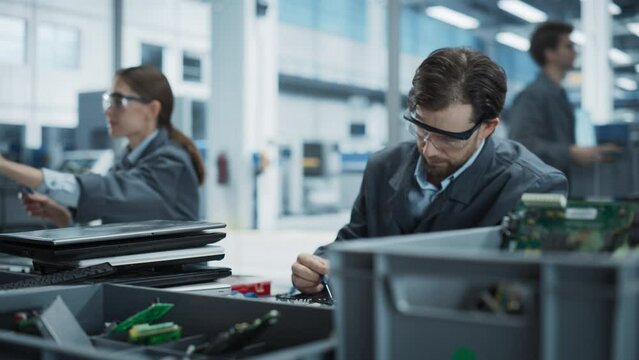 Caucasian Male And Female Workers Taking Apart Old Laptops To Recycle Electronic Components For Microchip Production At Modern Electronics Factory. Man And Woman Unscrewing and Sorting Computer Parts.