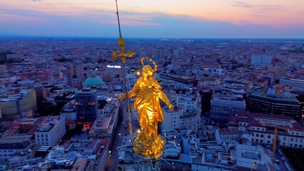 Aerial view of the statue of the Madonna on the central spire of the Duomo and the city's cathedral at dawn. Roofs of houses and skyscrapers. Sun over the horizon. duomo square. Italy Milan 16.11.2023 - obrazy, fototapety, plakaty