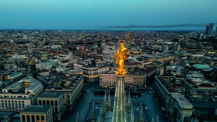 Aerial view of the statue of the Madonna on the central spire of the Duomo and the city's cathedral at dawn. Roofs of houses and skyscrapers. Sun over the horizon. duomo square. Italy Milan 16.11.2023 - obrazy, fototapety, plakaty