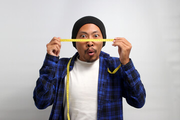 An enthusiastic young Asian man, clad in a beanie hat and casual shirt, gazes at a measuring tape with an expression of surprise or excitement, mouth slightly agape, against a white background