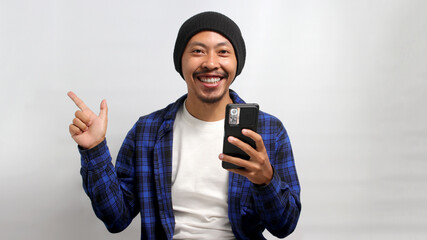 Excited Asian man, dressed in a beanie hat and casual shirt, points his finger at an empty space...
