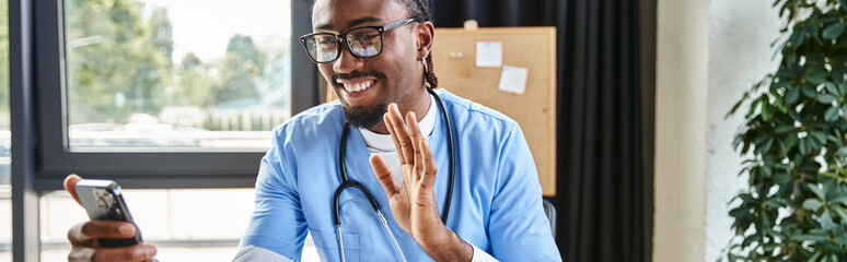 jolly african american doctor with glasses waving at phone camera and smiling joyfully, banner