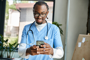 handsome happy african american doctor with stethoscope looking at his mobile phone, telemedicine - obrazy, fototapety, plakaty
