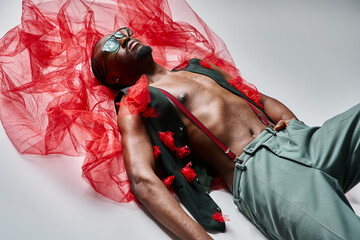 attractive young african american male model in stylish attire with red tulle fabric lying on floor