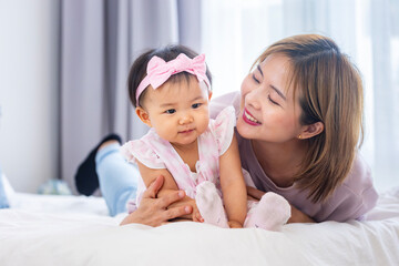 Asian mother is holding her pretty smiling baby daughter while spending quality time in bed for...
