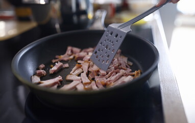 Close-up of female frying small slices of bacon on pan. Woman cooking tasty dinner or breakfast for family. Juicy and fresh meat with oil. Unhealthy nutrition concept