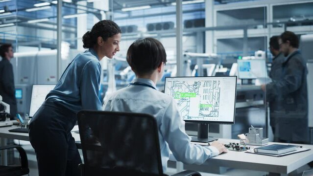 Hispanic Female Manager And Asian Male Engineer Using Desktop Computer With Microchip Blueprints at Automated Factory With Robotic Arms. Colleagues Discussing Production Hardware For Electronics.