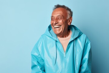 Obraz premium Portrait of a grinning indian elderly man in his 90s wearing a lightweight packable anorak against a pastel blue background. AI Generation