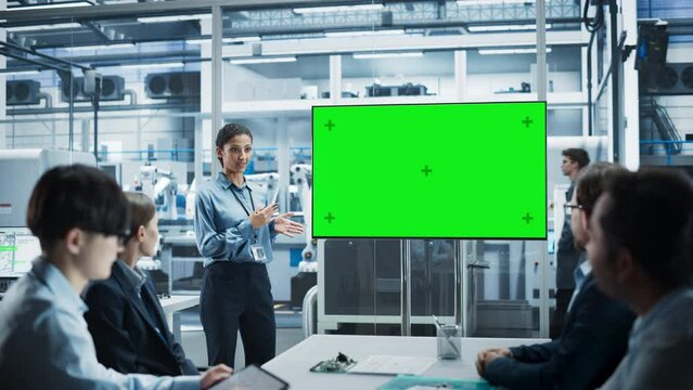 Autonomous Electronics Factory With Robotic Arms: Hispanic Female Team Lead Presenting Innovative Project To Diverse Engineers And Technicians On TV With Green Screen Chromakey In Office Meeting Room.