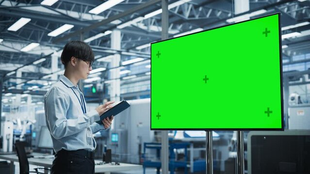 Asian Male Chief Engineer Using Tablet And Looking At TV With Green Screen Chromakey On Display At Autonomous Electronics Factory With Robotic Arms. Man Overlooking Assembly Of New Devices In Office.
