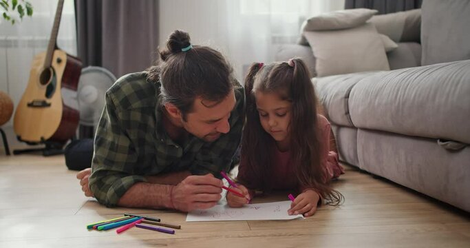 A little brunette girl in a pink dress lies on the floor and draws with her father, a brunette man in a checkered shirt, on a sheet of paper using multi-colored felt-tip pens in a modern apartment