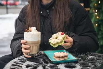 Foto op Canvas A woman drinks coffee with cream and eats Christmas macarons in a cafe outside. © puhimec