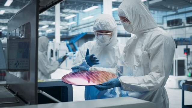 Semiconductor Production Process At Electronics Factory: Two Scientists In Sterile Suits Taking Out Silicon Wafer From Soldering Jet Printer And Checking It. Engineers Creating Hardware For Computers.