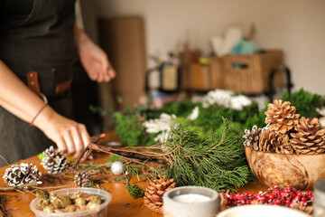 Workshop on making a Christmas wreath. A young woman collects a wreath of fir branches on a wooden table. DIY decorations. There are decorations for the wreath on the table: pine cones and ribbons.