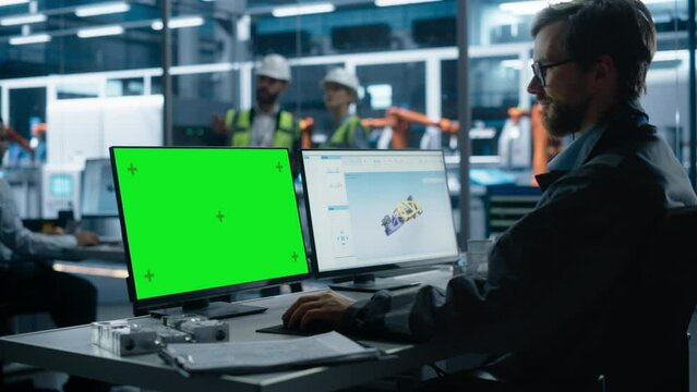 Caucasian Male Industrial Engineer Controlling Autonomous Conveyor With Robot Arms At Electric Engine Factory. Man Using Computer With Green Screen Chromakey On Display To Adjust Automated Production.