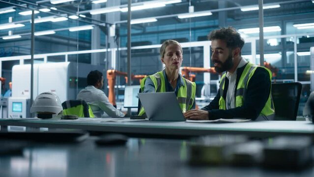 Autonomous Factory Office: Caucasian Female Production Supervisor Talks to Hispanic Male Process Engineer Working on Laptop Computer. Automated Robot Arm Assembly Line Manufacturing Machinery.