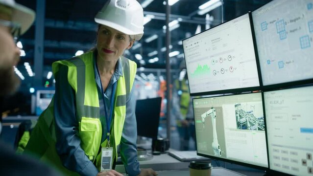 Caucasian Male Process Engineer Using Multi Monitor Workstation And Talking To Female Quality Control Technician At Electronics Factory. Colleagues Monitoring Autonomous Assembly Line With Robot Arms.