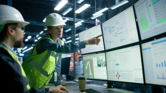 Caucasian Male Process Engineer Using Multi Monitor Workstation And Talking To Female Supervisor At Industrial Machinery Factory. Colleagues Monitoring Autonomous Assembly Line With Robotic Arms.