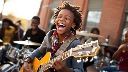 Foto op Plexiglas A young Black boy joyfully plays a yellow acoustic guitar outdoors, while other children play percussion instruments in the background. © volga