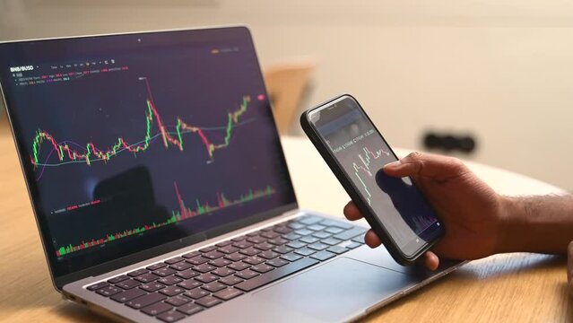 Crypto investor looking at smartphone and laptop screens, using cell phone app, executing financial stock trade market trading order to buy or sell cryptocurrency, trader working remotely