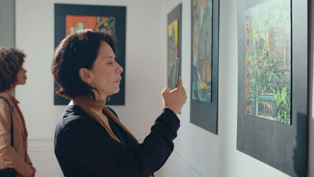 Interested mid-aged woman observing collection in art gallery and photographing painting with mobile phone