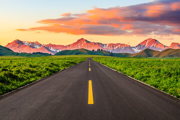 Asphalt road and green meadow with mountain nature landscape at sunset