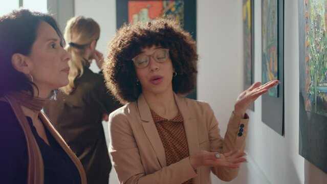 Female gallery guide discussing artwork wit mid-aged female visitor looking attentively at painting and asking questions about masterpiece