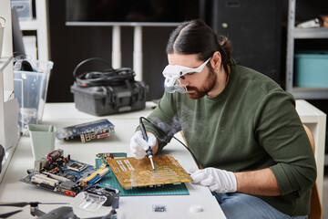 Side view portrait of bearded repairman fixing circuit board with soldering iron in small tech...
