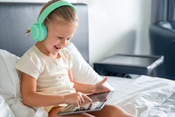 Happy little girl in headphones using digital tablet and smiling happy while listening to music or...