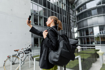 Side view of fashionable and fair haired young woman with backpack taking selfie on smartphone