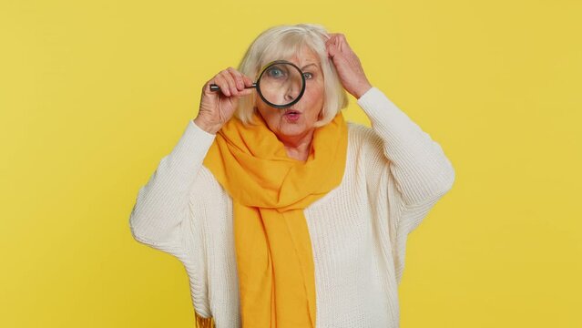 Investigator researcher scientist senior old woman holding magnifying glass near face, looking into camera with big zoomed funny eye, searching, analyzing. Elderly grandmother on yellow background