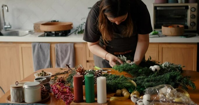 A young woman at home in her kitchen prepares for the Christmas celebration and makes a wreath from fir branches on a wooden table. Homely atmosphere of preparation for the New Year.