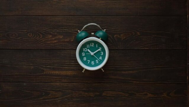 Female hand puts classic green alarm clock on brown wooden background, top view