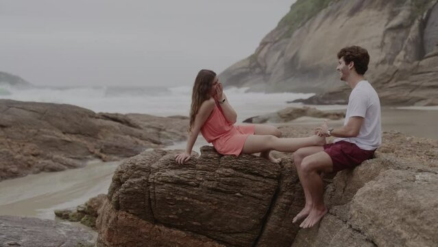 Homem jovem massageando o pe de sua namorada, enquanto conversam sentados em uma rocha na praia. Cinematico 4k.
