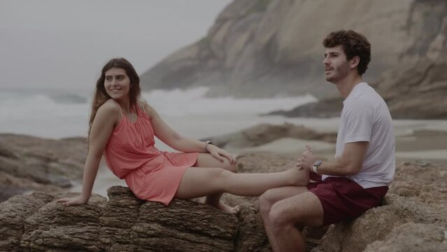 Homem jovem massageando o p&eacute; de sua namorada, enquanto conversam sentados em uma rocha na praia. Cinematico 4k.