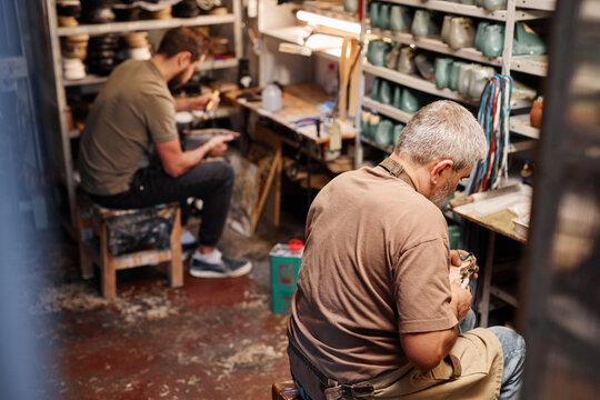 Mature man creating or repairing shoes while sitting in front of camera by shelves with workpieces and young male apprentice on background