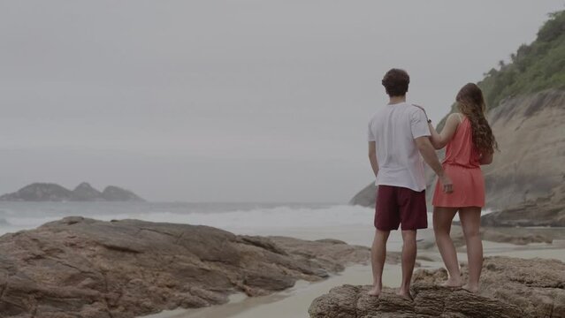 Casal jovem sorridente, se divertindo, conversando, em pe sobre uma pedra na praia. Cinematico 4k.