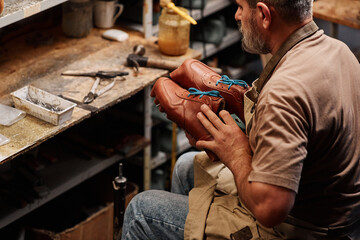 Side view of mature professional shoemaker holding brown leather workpieces while sitting in front...