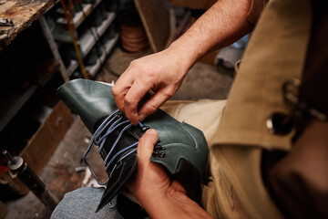 Hands of professional male shoemaker putting shoelace into upper part of unfinished black leather boot while sitting in workshop - obrazy, fototapety, plakaty