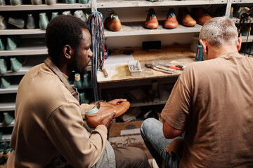 Young African American man in workwear looking at shoemaking master while sitting next to him in...