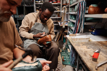 Young African American man in apron putting together upper part of footwear with sole while working...