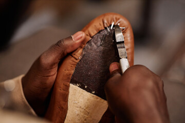 Above angle of hands of young black man fixing leather with nails and pliers on sole of boot or...