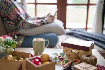 Young asian woman surfing social media on smartphone and cup of hot chocolate with marshmallow on table while sitting on couch in living room decorating xmas ornaments for celebrate christmas festive
