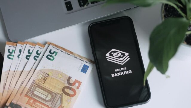 A smartphone with an image of online banking lies on a table with money in cash. Advantage of online banking. A hand puts a smartphone on top of money.