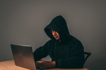A male hacker wearing glasses and a hoodie covers his head in the dark is hacking into a victim's...