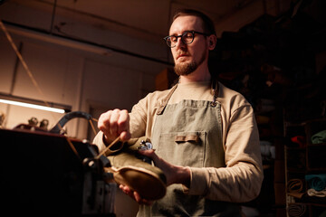 Young male manufacturer stretching thread in sewing machine while using electric equipment in process of creating new footwear